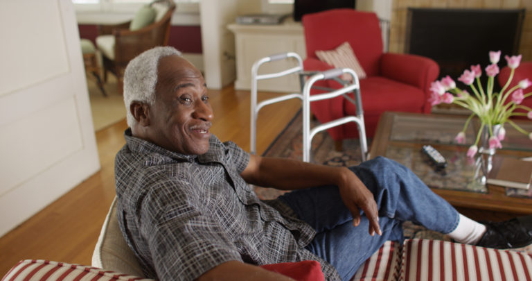 A senior man sitting on a couch with a walker next to him.